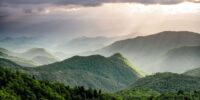 Sunlight filters through the clouds, casting a dramatic light on the lush, rolling hills below. The shades of green and the depth of the valleys create a sense of serene complexity, as nature's grandeur is showcased under a stormy sky along the Blue Ridge Parkway in North Carolina.