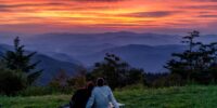 A couple sits closely together on a blanket, gazing at a breathtaking sunset over layered mountain ridges. The sky is ablaze with shades of orange, pink, and purple, contrasting with the darkened silhouettes of the rolling hills.