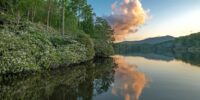 A tranquil Price Lake reflects the vibrant colors of a cloud illuminated by the sunset, while the surrounding foliage casts reflections on the water. The stillness of the water provides a mirror image of the scenery, enhancing the serene atmosphere of the landscape. Blue Ridge Parkway, North Carolina.