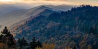 Rolling hills covered in trees displaying autumn colors gently recede into the distance under a sky streaked with clouds. The rising sun casts a warm light over the landscape, highlighting the layers of the mountains and the misty horizon. Great Smoky Mountains National Park.