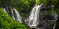 At Soco Falls in North Carolina, a cascade of water tumbles down a rocky incline surrounded by lush green foliage. The scene captures the serenity and power of a natural waterfall nestled within a dense forest.