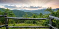 In the Great Smoky Mountains National Park, a serene sunset bathes the landscape in a warm glow, silhouetting a rustic wooden fence in the foreground against a backdrop of rolling hills. The sky above is a vibrant tableau of colors, with sun rays piercing through the clouds to illuminate the lush greenery of the valley below.